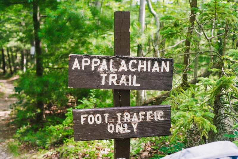 The image shows a wooden signpost in a wooded area. The top sign reads "Appalachian Trail" and the bottom sign reads "Foot Traffic Only." The signs are weathered and appear to be located at the entrance to a hiking trail. The surrounding vegetation is lush and green, suggesting a natural environment.